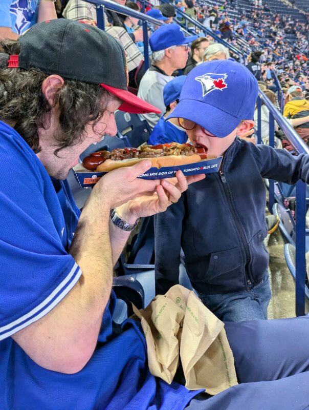 Padre e hijo comiendose un perrito caliente enorme durante un partido de los Blue Jays de Toronto.